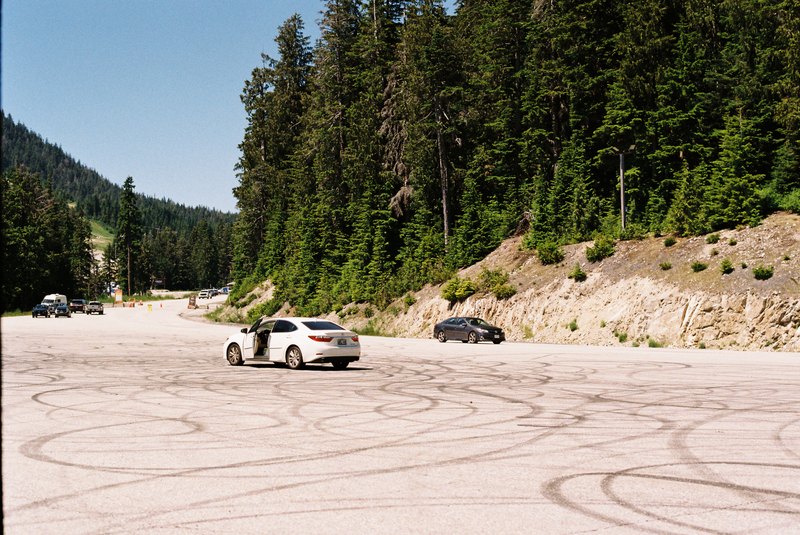 A white car with an open door in a mountain parking lot marked with tire tracks.