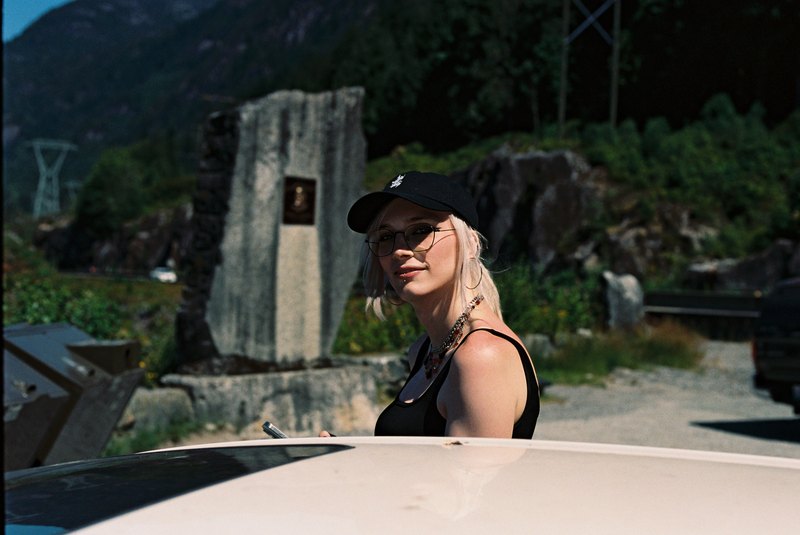 A person wearing a black cap and glasses beside a car in the mountains.