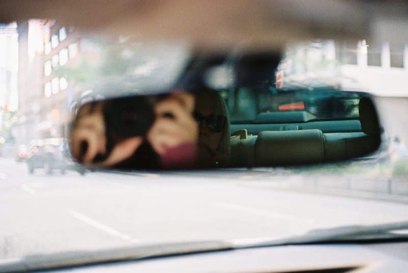 A camera and its photographer reflected in a car rearview mirror.